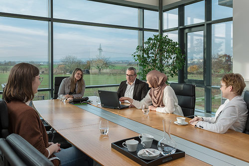Frauen und Männer am Konferenztisch vor großen Fenstern mit Blick in die Natur.