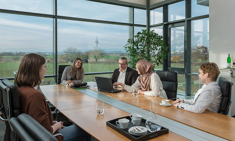 Frauen und Männer am Konferenztisch vor großen Fenstern mit Blick in die Natur.